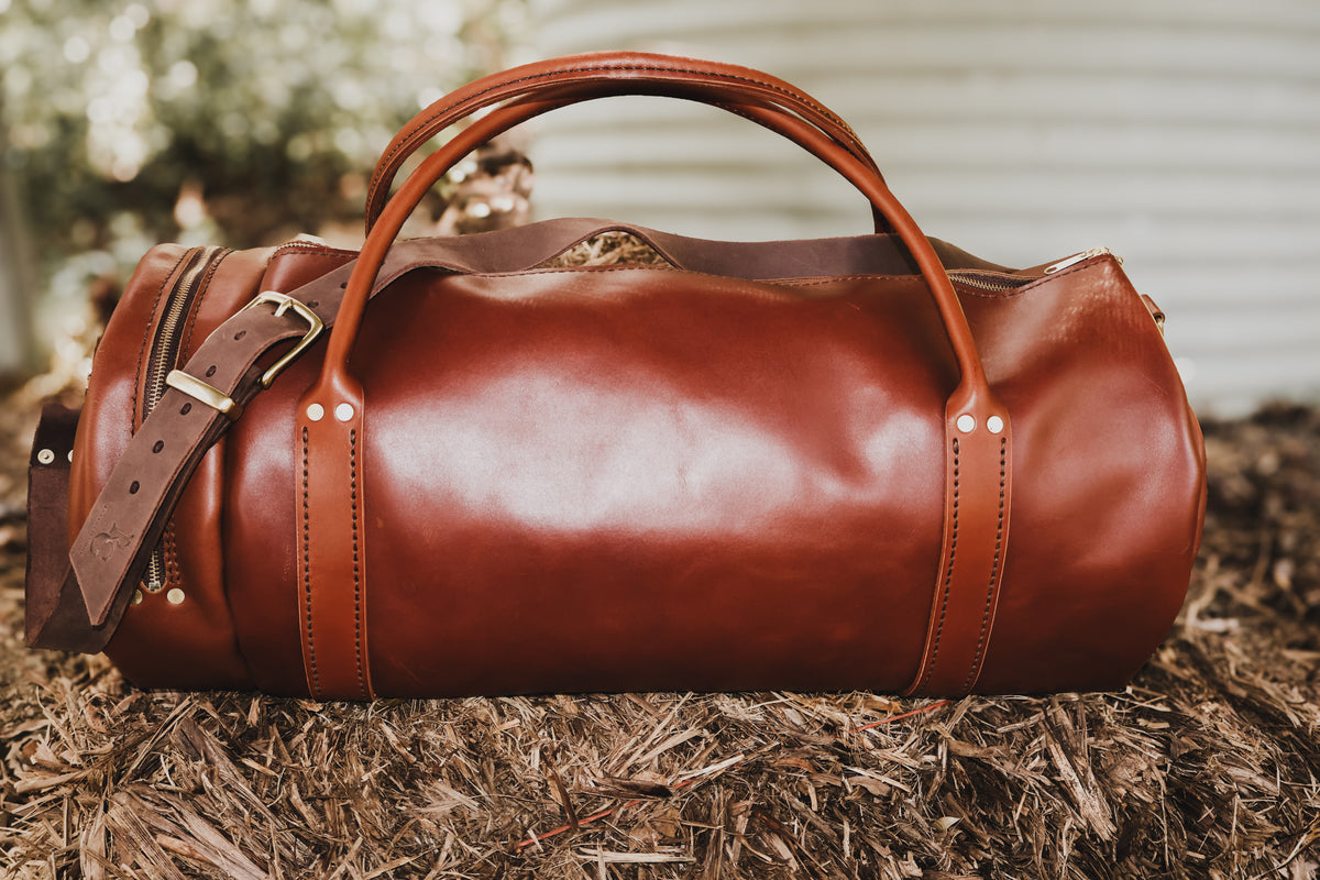 Side view of The Bully chestnut full-grain leather duffel bag outdoors, showing its rolled handles, shoulder strap and brass hardware.